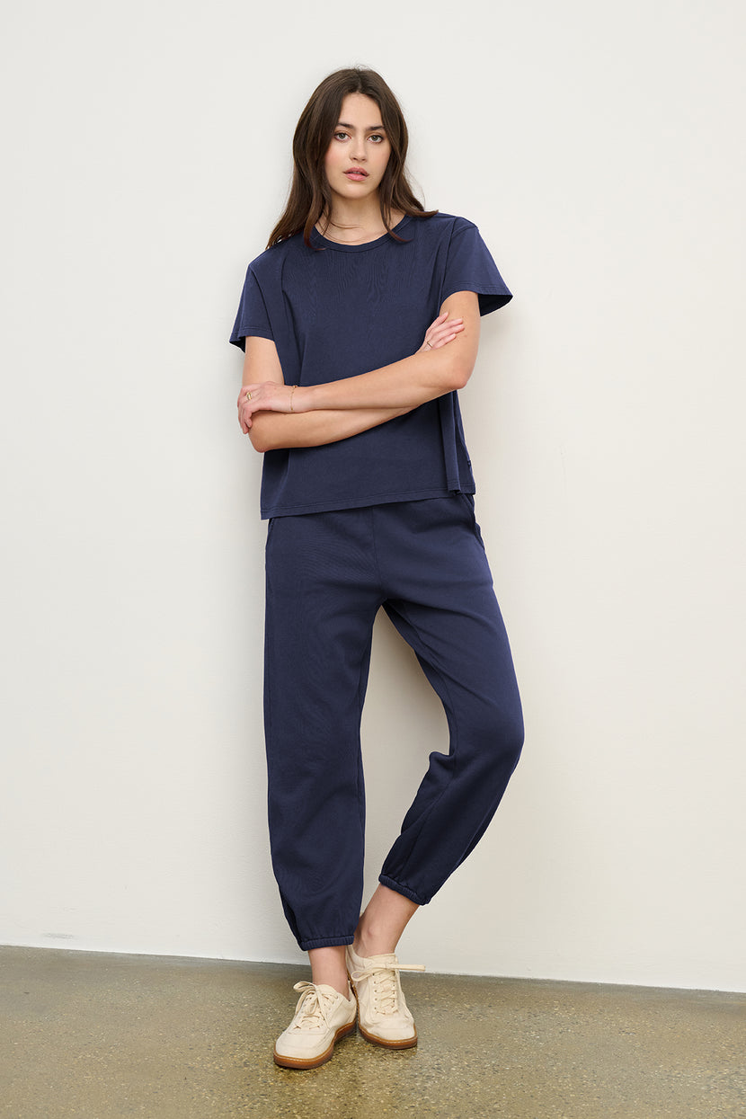 A woman stands against a white wall wearing the Velvet by Jenny Graham ZUMA SWEATPANT in navy with a matching short-sleeve top and beige sneakers.