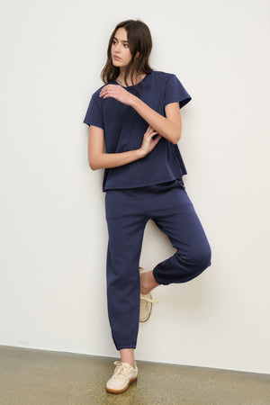 A woman stands against a white wall wearing a navy blue short-sleeve top and matching ZARA SWEATPANT by Velvet by Graham & Spencer in a relaxed fit, paired with beige shoes.
