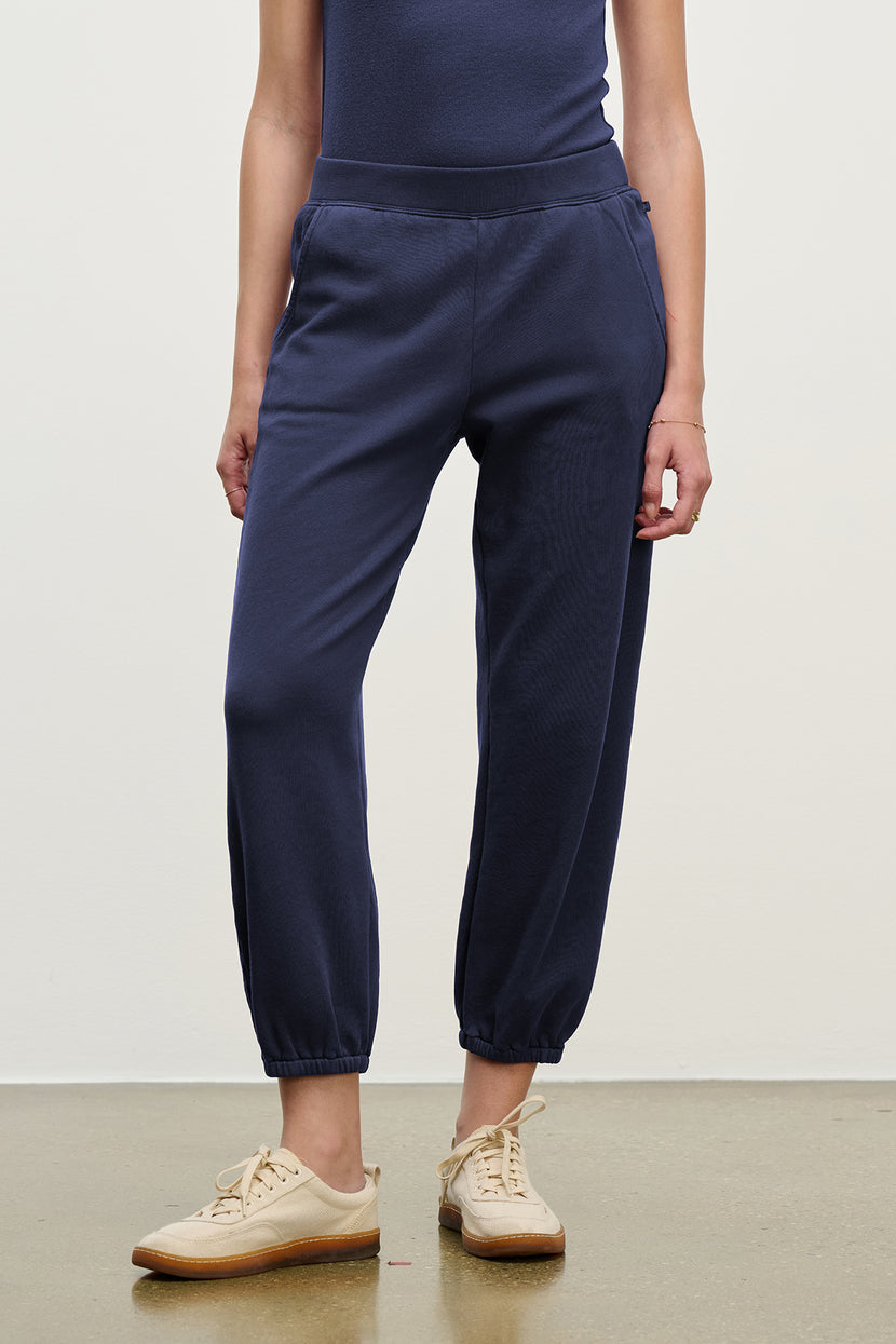Dressed in Velvet by Jenny Graham's ZUMA SWEATPANT in navy blue with a matching top and beige sneakers, a person stands against a plain background.