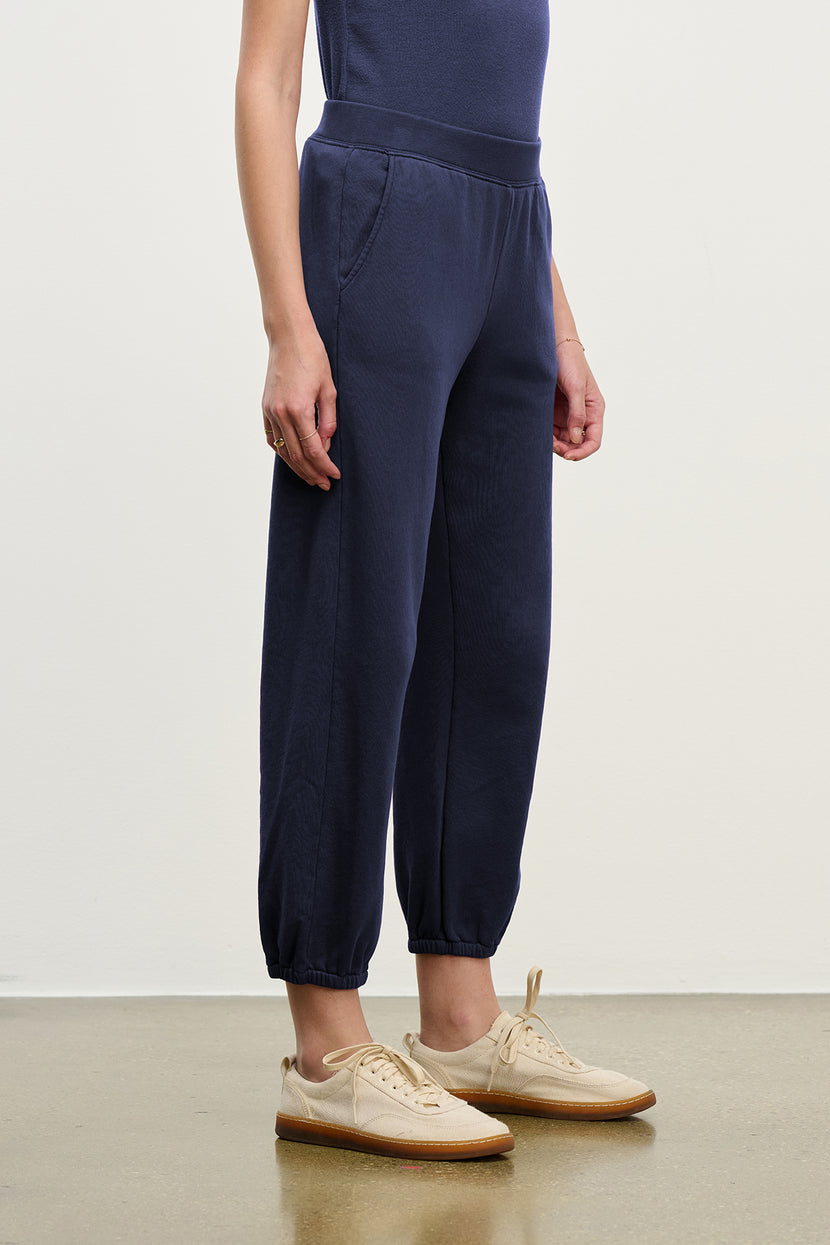 A person stands indoors wearing the ZUMA SWEATPANT by Velvet by Jenny Graham in navy blue organic cotton, with a matching top and beige sneakers, against a plain background.