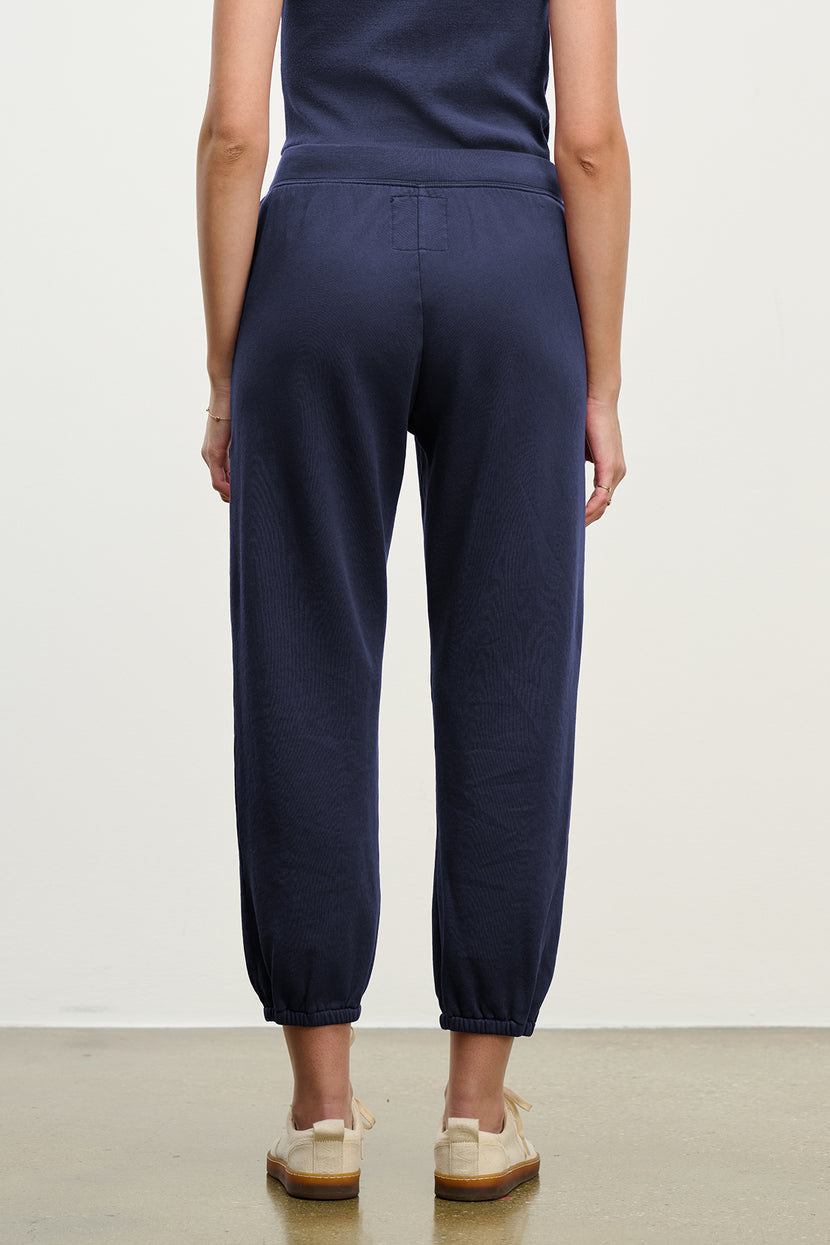 A person stands indoors facing away, wearing Velvet by Jenny Graham's ZUMA SWEATPANT in navy blue with a matching top and beige shoes.