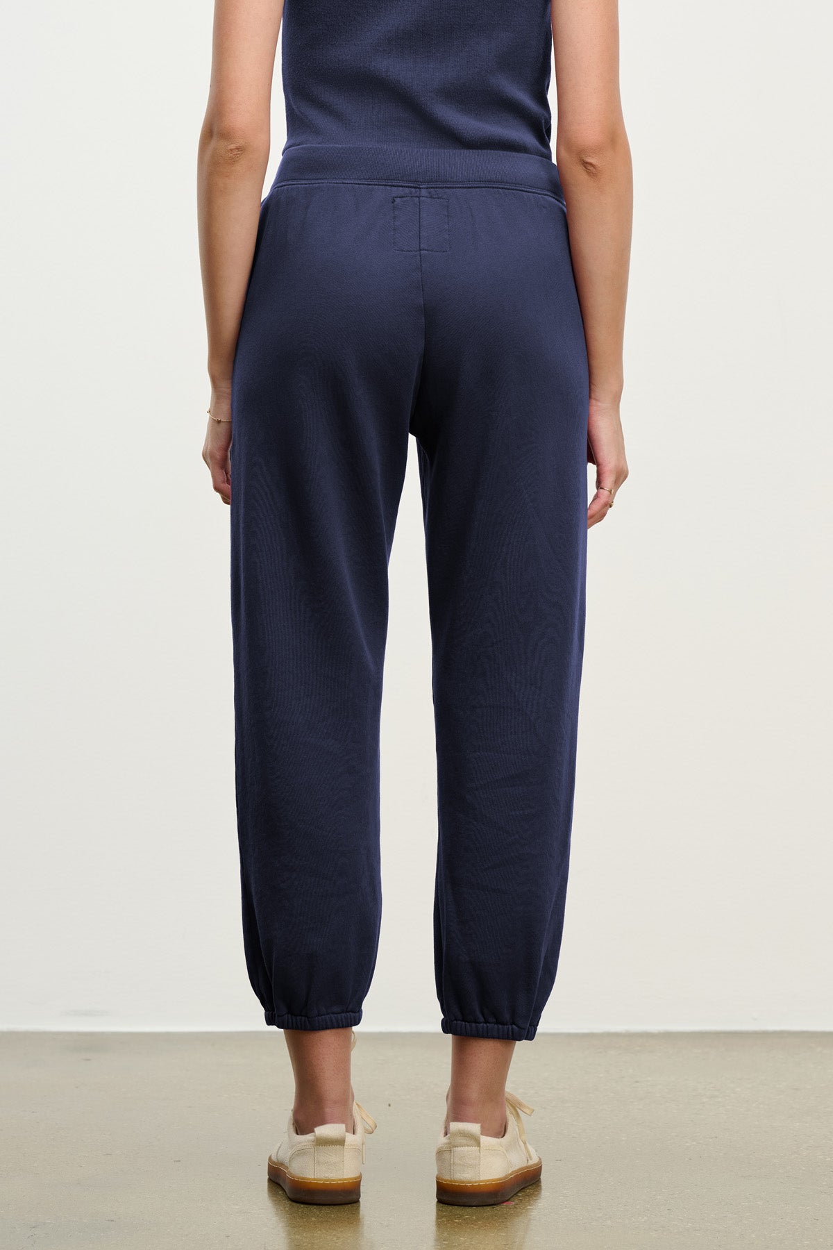 A person stands facing away, wearing Velvet by Graham & Spencer ZARA SWEATPANT joggers in navy blue cotton fleece and beige shoes, arms relaxed at their sides against a plain background.-40294545490113