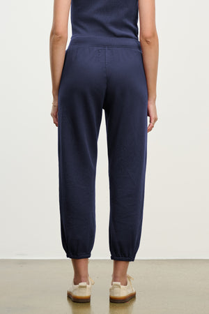 A person stands facing away, wearing Velvet by Graham & Spencer ZARA SWEATPANT joggers in navy blue cotton fleece and beige shoes, arms relaxed at their sides against a plain background.