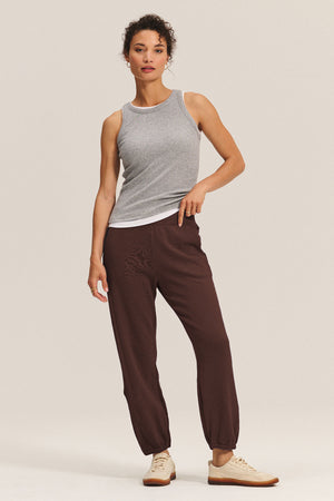 A woman stands against a plain background wearing a grey sleeveless tank top, Velvet by Graham & Spencer ZARA SWEATPANT in a relaxed fit, and beige sneakers.