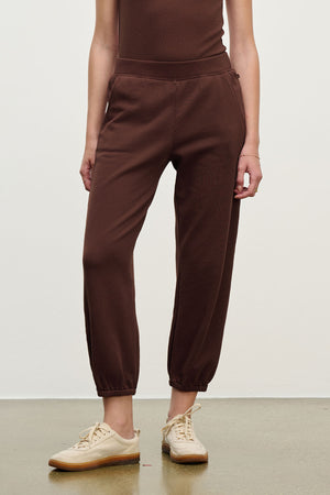 A person wearing relaxed fit ZARA SWEATPANT joggers by Velvet by Graham & Spencer in brown, paired with beige sneakers, stands against a plain light background.