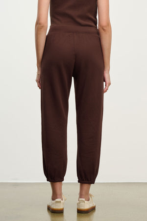 A person is standing facing away from the camera against a plain background, wearing relaxed fit brown ZARA SWEATPANT joggers by Velvet by Graham & Spencer and beige slip-on shoes.