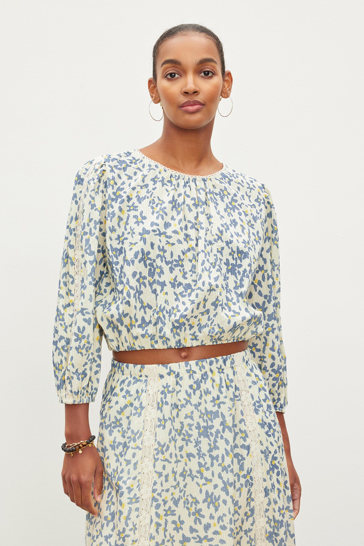   A woman stands against a plain background wearing a light-colored, floral-patterned CAMERON TOP by Velvet by Graham & Spencer with three-quarter sleeves and a matching skirt. She has short hair and is wearing hoop earrings and a bracelet. 