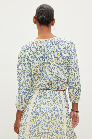 Back view of a person with a bun hairstyle, wearing hoop earrings and a patterned outfit consisting of the CAMERON TOP by Velvet by Graham & Spencer and a skirt in light colors with blue and yellow designs.