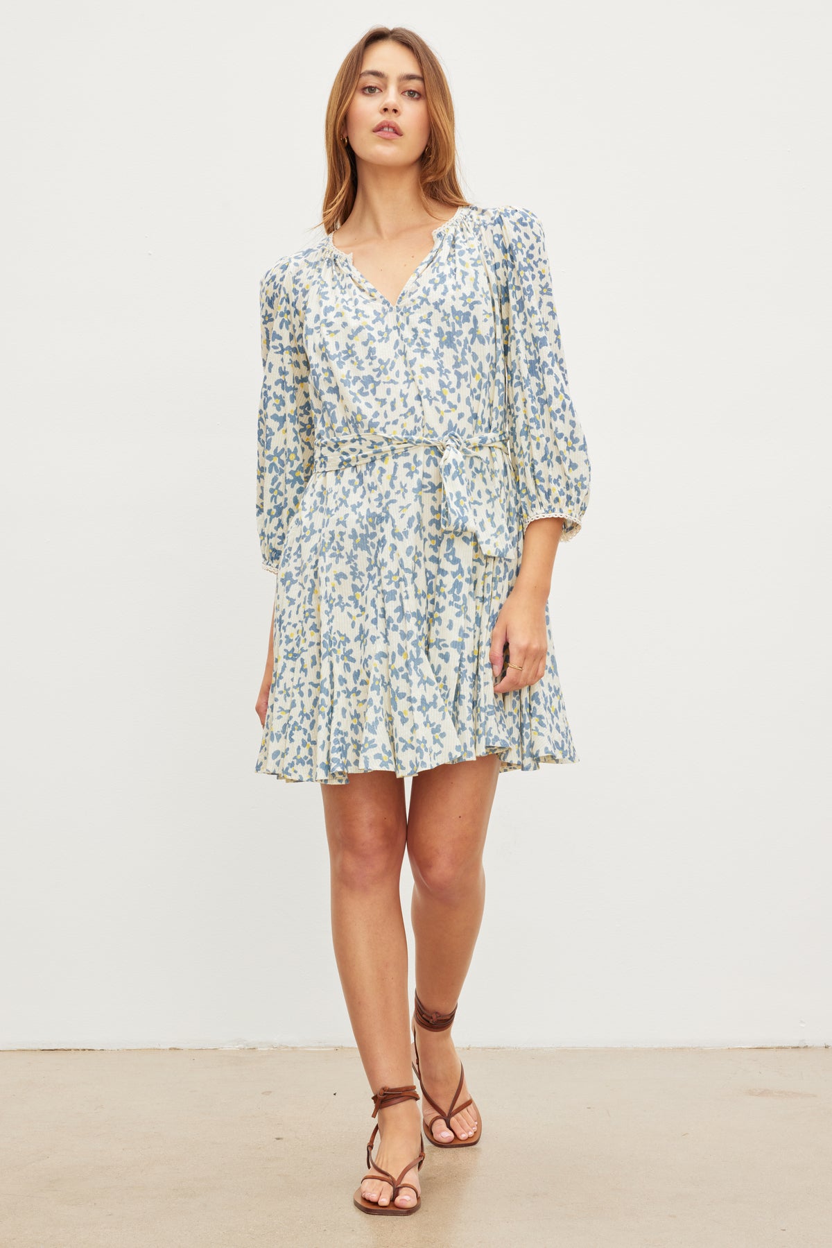   A woman stands against a plain white background wearing the MARIAH DRESS by Velvet by Graham & Spencer, a light-colored, floral print wrap dress with long sleeves, and brown strappy sandals. 