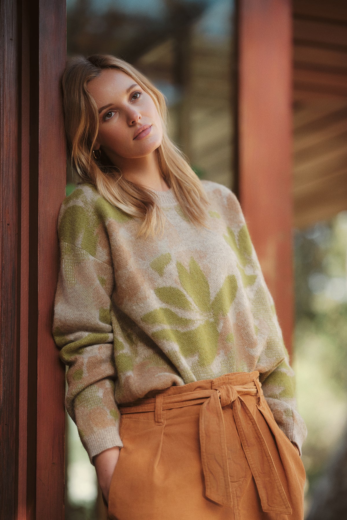   A woman with long blonde hair leans against a wooden surface. She wears the Velvet by Graham & Spencer RIVA SWEATER, a cozy light piece featuring a green leaf pattern, and high-waisted tan trousers tied with a belt. 