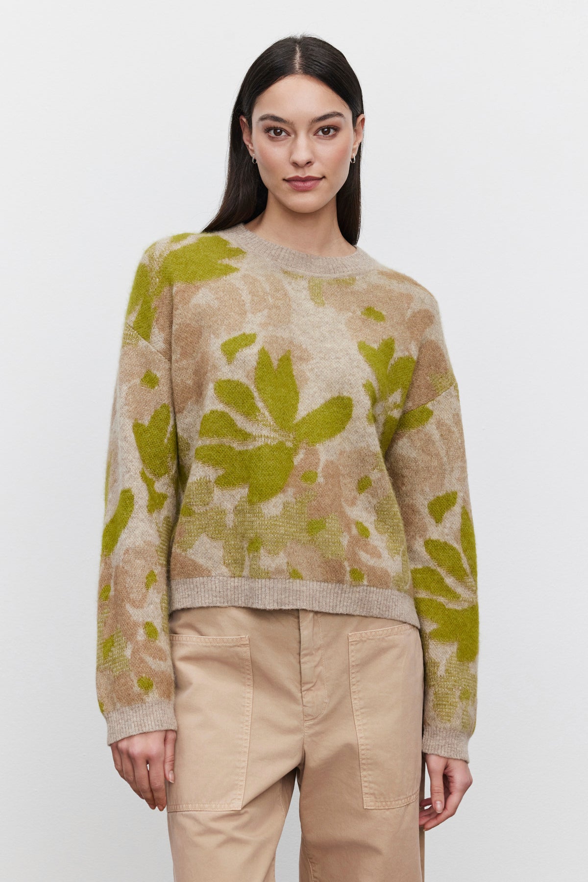   A person with long dark hair wears a beige and green floral jacquard patterned RIVA SWEATER from Velvet by Graham & Spencer and beige pants, standing against a plain white background. 