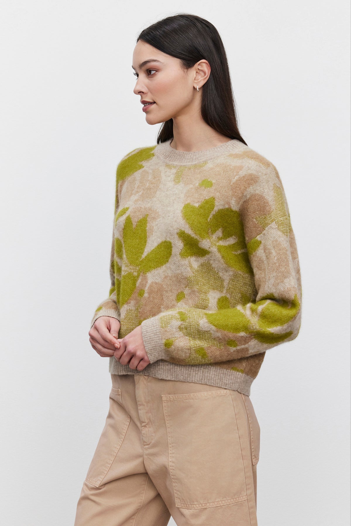   A woman with long dark hair wears the RIVA SWEATER from Velvet by Graham & Spencer, featuring a beige and green floral jacquard pattern, along with beige pants, standing against a plain white background. 