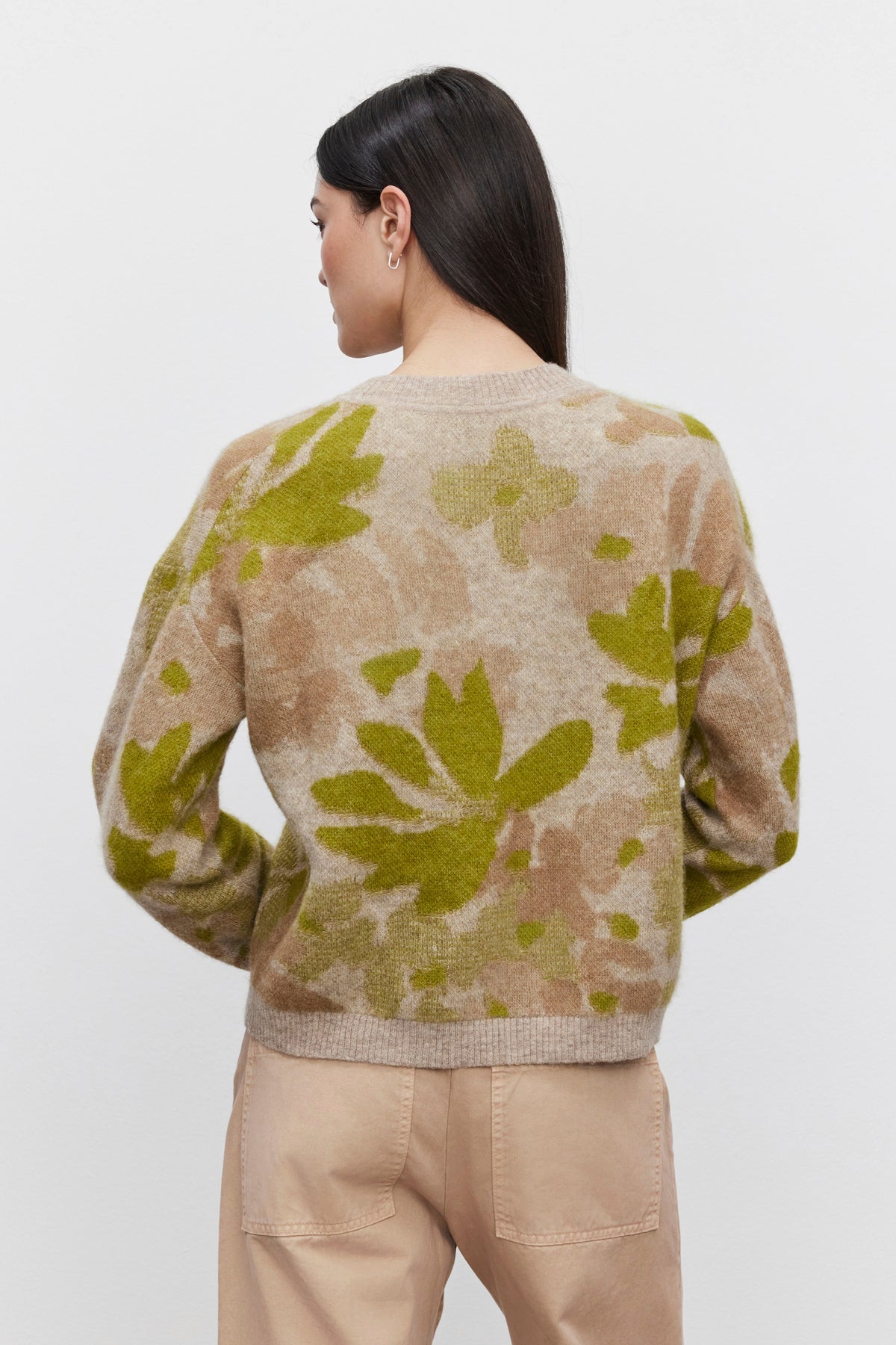   A woman with long, dark hair is facing away, wearing a beige RIVA SWEATER by Velvet by Graham & Spencer with green and brown floral jacquard patterns and light beige pants. 