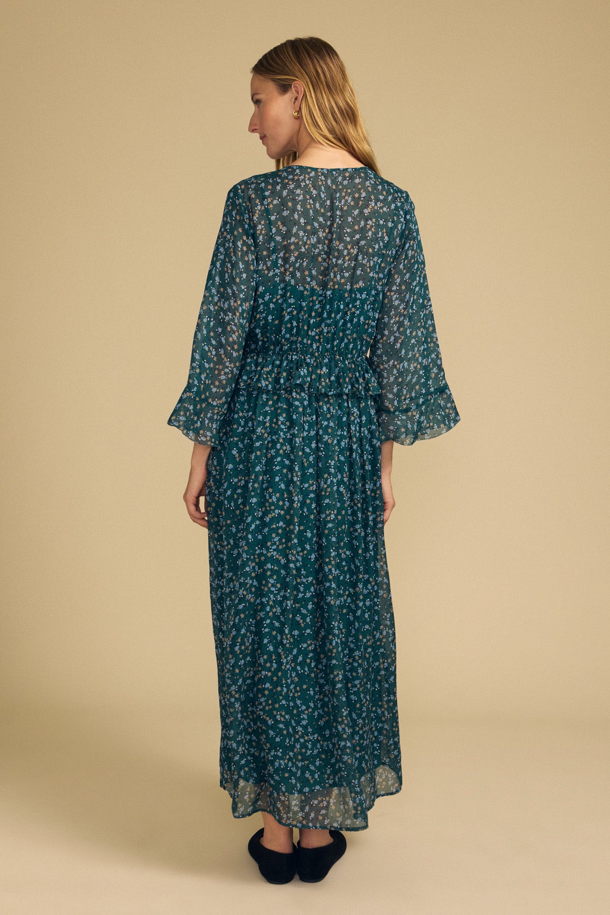 A woman stands facing away, wearing the ISABELA DRESS by Velvet by Graham & Spencer—a long teal dress with a floral pattern and loose sleeves. The background is plain beige.-41826786738369
