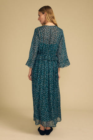 A woman stands facing away, wearing the ISABELA DRESS by Velvet by Graham & Spencer—a long teal dress with a floral pattern and loose sleeves. The background is plain beige.