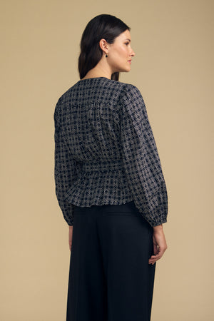 A woman stands sideways and facing away against a plain beige background, wearing the Velvet by Graham & Spencer IMANI BLOUSE—a long-sleeve, patterned blouse—paired with black pants.