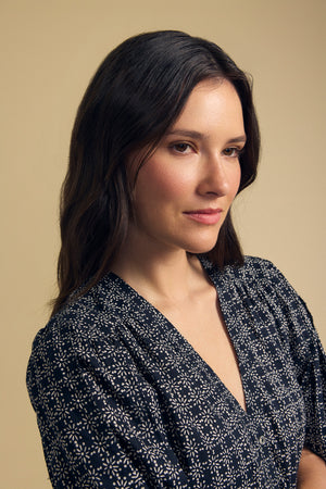 A woman with long brown hair wears the IMANI BLOUSE by Velvet by Graham & Spencer and looks to the side against a plain beige background.
