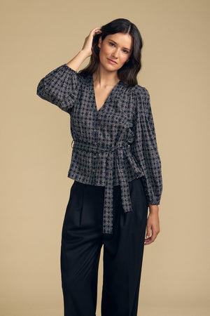 A woman stands against a beige background wearing the IMANI BLOUSE by Velvet by Graham & Spencer—a patterned wrap blouse with a tie—paired with black wide-leg pants. She touches her hair with one hand and looks at the camera.
