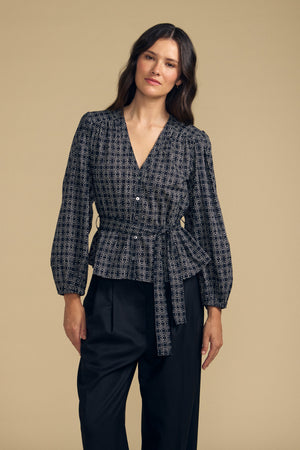 A woman wearing the Velvet by Graham & Spencer IMANI BLOUSE—a patterned black top with a waist tie—paired with black pants, standing against a plain beige background.