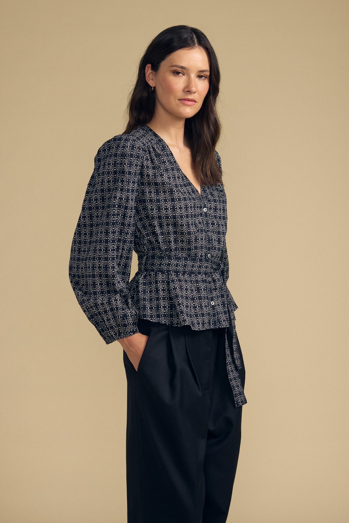 A woman stands against a beige background wearing the IMANI BLOUSE by Velvet by Graham & Spencer—a dark patterned, long-sleeve top—paired with black high-waisted trousers and her hands in her pockets.-41826786508993
