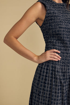 A person wearing the KATHERINE DRESS by Velvet by Graham & Spencer—a sleeveless, textured navy cotton dress with a white pattern—stands with one hand on their hip against a beige background.