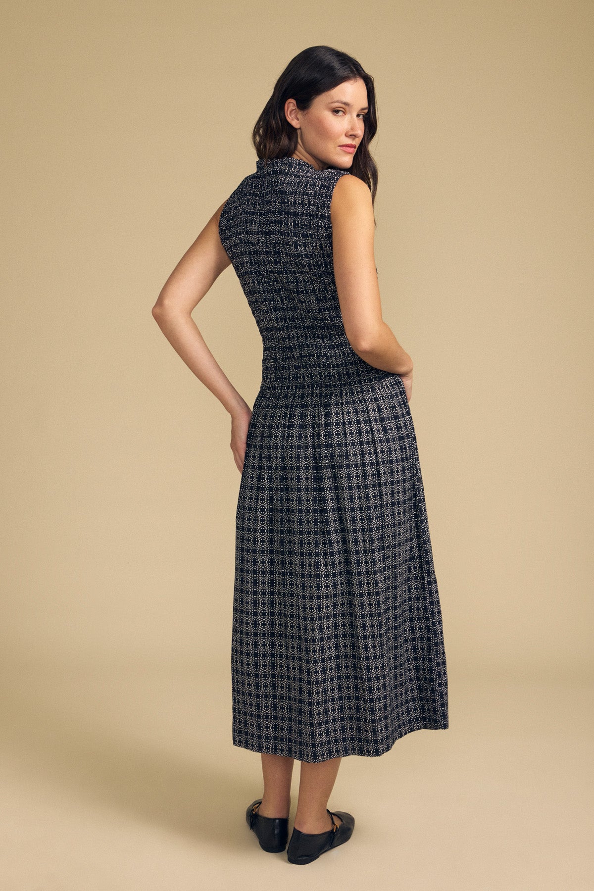 A woman stands on a beige background, facing slightly away from the camera, wearing the KATHERINE DRESS by Velvet by Graham & Spencer—a sleeveless, ankle-length black and gray patterned cotton dress—paired with black flat shoes.-41834062184641
