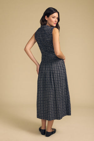 A woman stands on a beige background, facing slightly away from the camera, wearing the KATHERINE DRESS by Velvet by Graham & Spencer—a sleeveless, ankle-length black and gray patterned cotton dress—paired with black flat shoes.