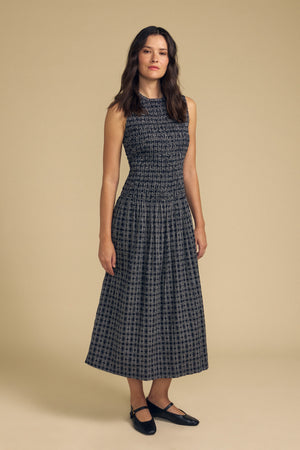 A woman stands against a beige background wearing the KATHERINE DRESS by Velvet by Graham & Spencer—a sleeveless, dark patterned cotton midi dress—paired with black flat shoes and her hair down.