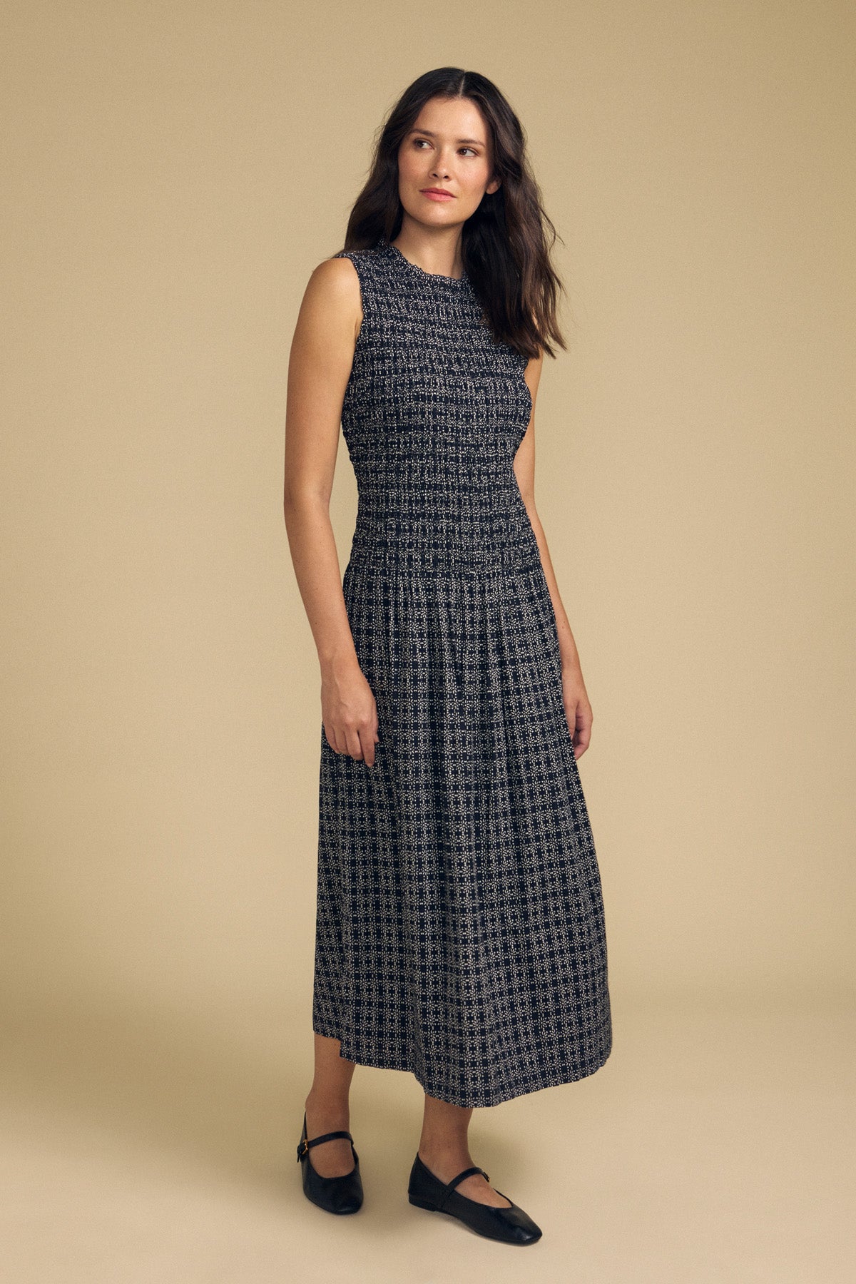 Woman standing against a beige background, wearing the KATHERINE DRESS by Velvet by Graham & Spencer—a sleeveless, ankle-length black cotton dress with subtle patterns—paired with black flat shoes. Her hands rest relaxed by her sides as she gazes slightly to her left, making this dress the perfect versatile choice for summer.-41834062119105