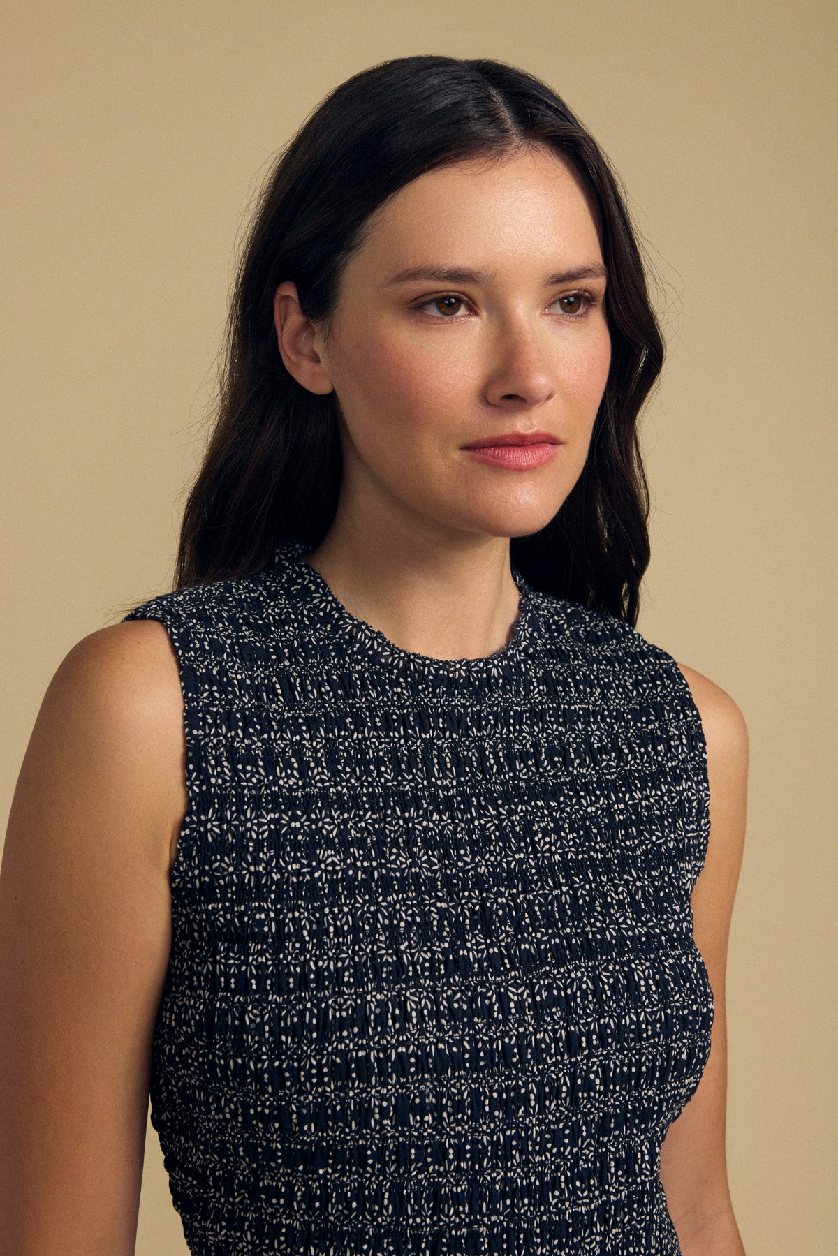 A woman with long dark hair stands against a plain beige background, wearing the KATHERINE DRESS by Velvet by Graham & Spencer—a sleeveless, textured black and white piece that captures effortless summer elegance as she gazes softly to the side.