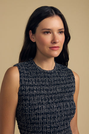 A woman with long dark hair stands against a plain beige background, wearing the KATHERINE DRESS by Velvet by Graham & Spencer—a sleeveless, textured black and white piece that captures effortless summer elegance as she gazes softly to the side.