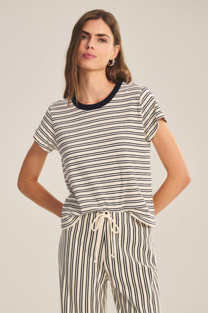 A woman wears the Velvet by Graham & Spencer FOSTER TEE—a short-sleeved, black and white striped jersey top with a contrast crew neckline—standing with one hand on her hip against a plain background.