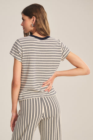 A woman faces away from the camera wearing the Velvet by Graham & Spencer FOSTER TEE, a cream and black striped cropped jersey top with matching pants and a contrast crew neckline.
