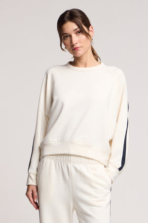 A woman wears the FRANKIE SWEATSHIRT by Velvet by Graham & Spencer— a midweight fleece top with black sleeve stripes—paired with matching cream sweatpants for a coordinated loungewear look against a plain background.