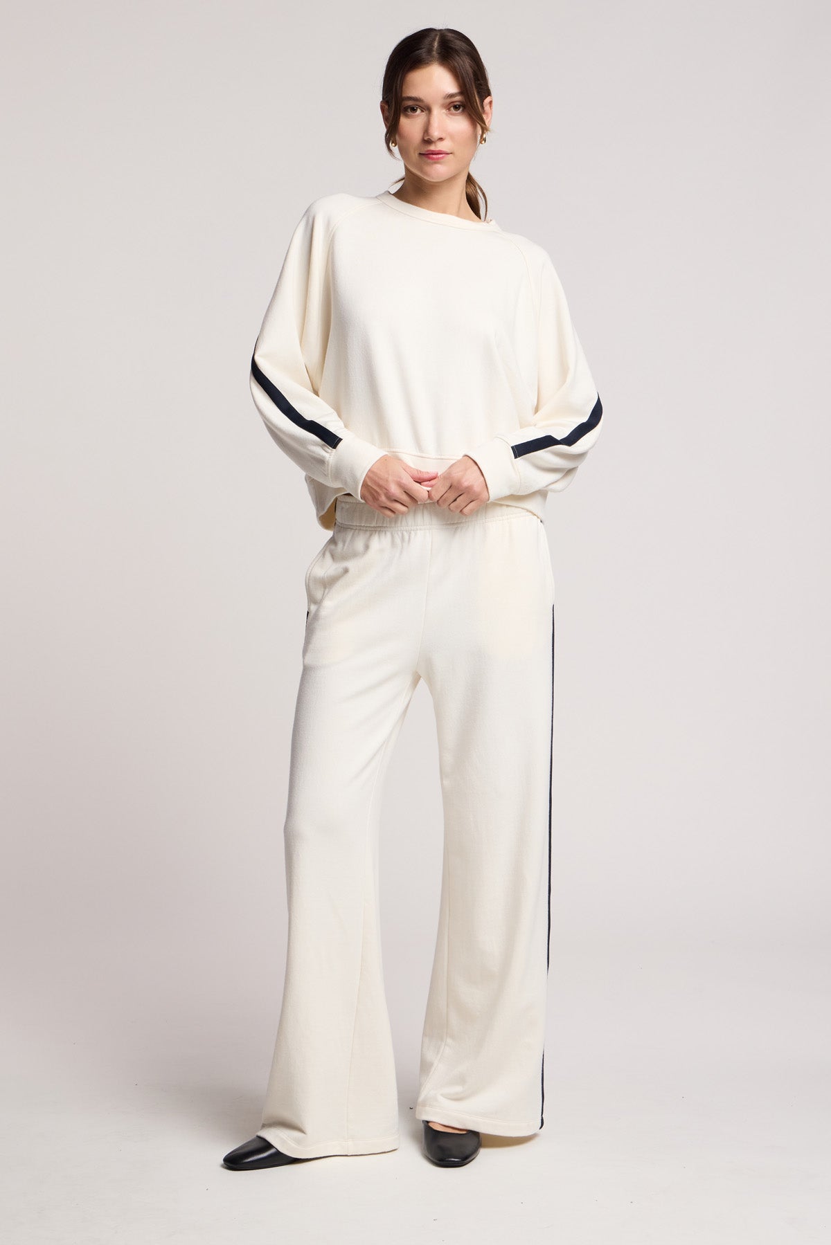 A woman stands in a studio wearing the FRANKIE SWEATSHIRT by Velvet by Graham & Spencer—an off-white lounge set with black sleeve and pant stripes, hands in pockets, paired with black pointed shoes.