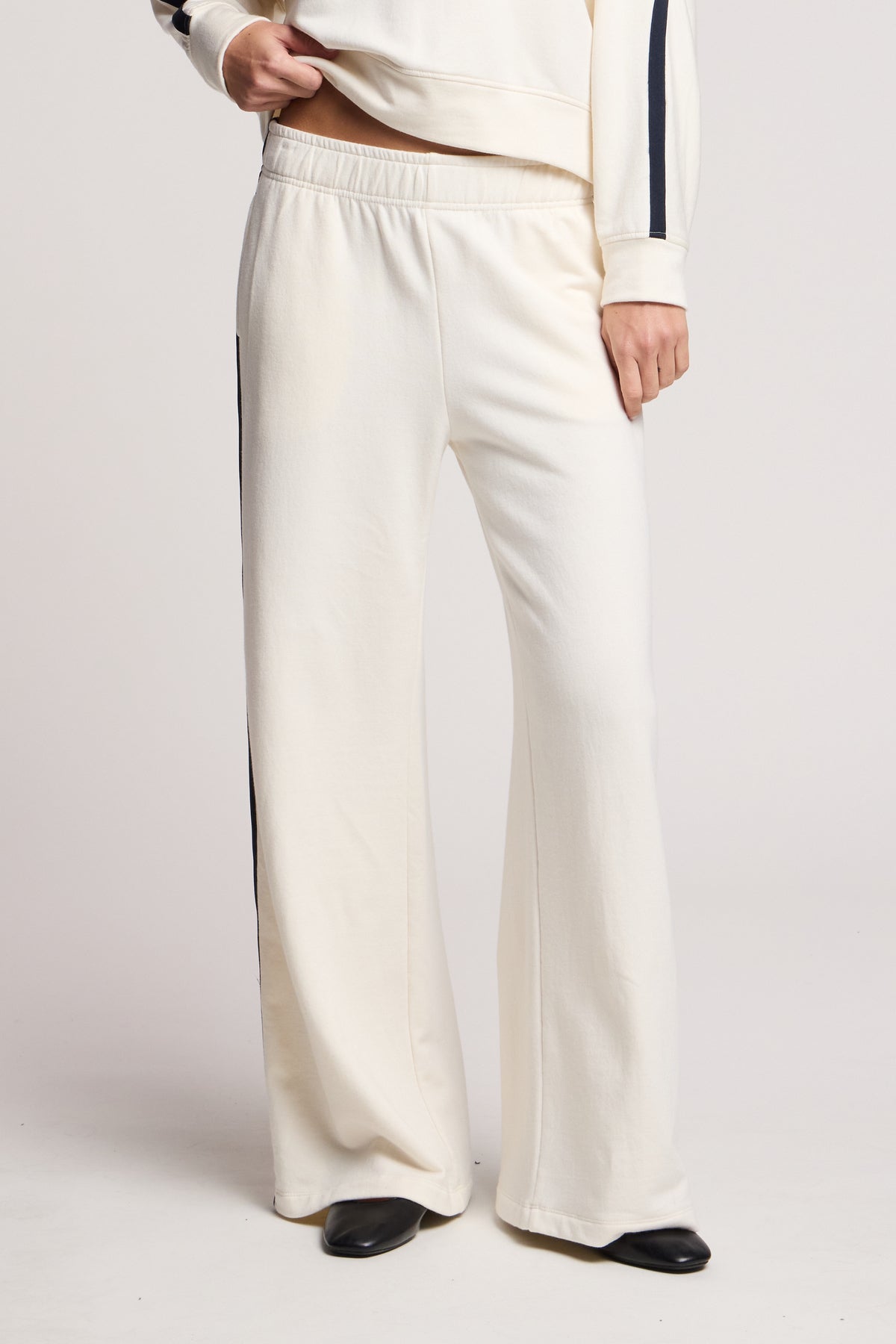 A person wears the Velvet by Graham & Spencer GIGI SWEATPANT—cream, wide-leg fleece pants with black side stripes and an elastic waist—styled with a matching cream top and black shoes.