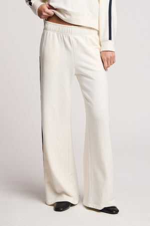 A person wears the Velvet by Graham & Spencer GIGI SWEATPANT—cream, wide-leg fleece pants with black side stripes and an elastic waist—styled with a matching cream top and black shoes.