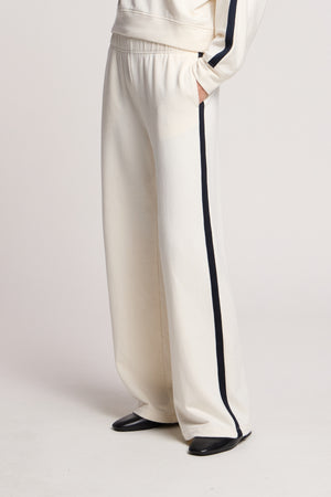 Model wears the GIGI SWEATPANT by Velvet by Graham & Spencer, featuring a relaxed fit, cream color, wide legs, and black side stripes with black shoes against a plain background.