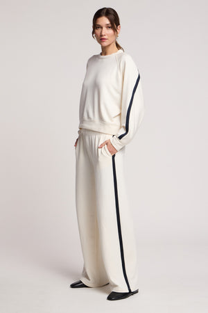 A woman wears the GIGI SWEATPANT by Velvet by Graham & Spencer—an off-white, wide-leg lounge set with black sleeve stripes and elastic waist, hands in pockets, posed against a plain light background.