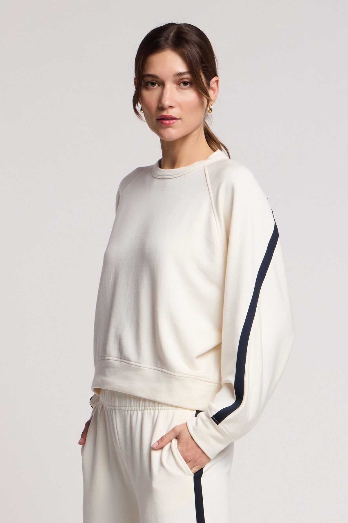 A woman wears the FRANKIE SWEATSHIRT by Velvet by Graham & Spencer, a cream-colored top with black sleeve stripes, paired with matching pants, showcasing chic coordinated loungewear against a plain background.-40732603318465