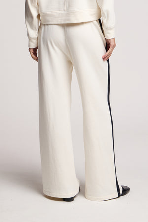 Model wears the GIGI SWEATPANT by Velvet by Graham & Spencer—loose-fitting, off-white wide-leg pants with black side stripes and elastic waist—shown from the back against a plain background.