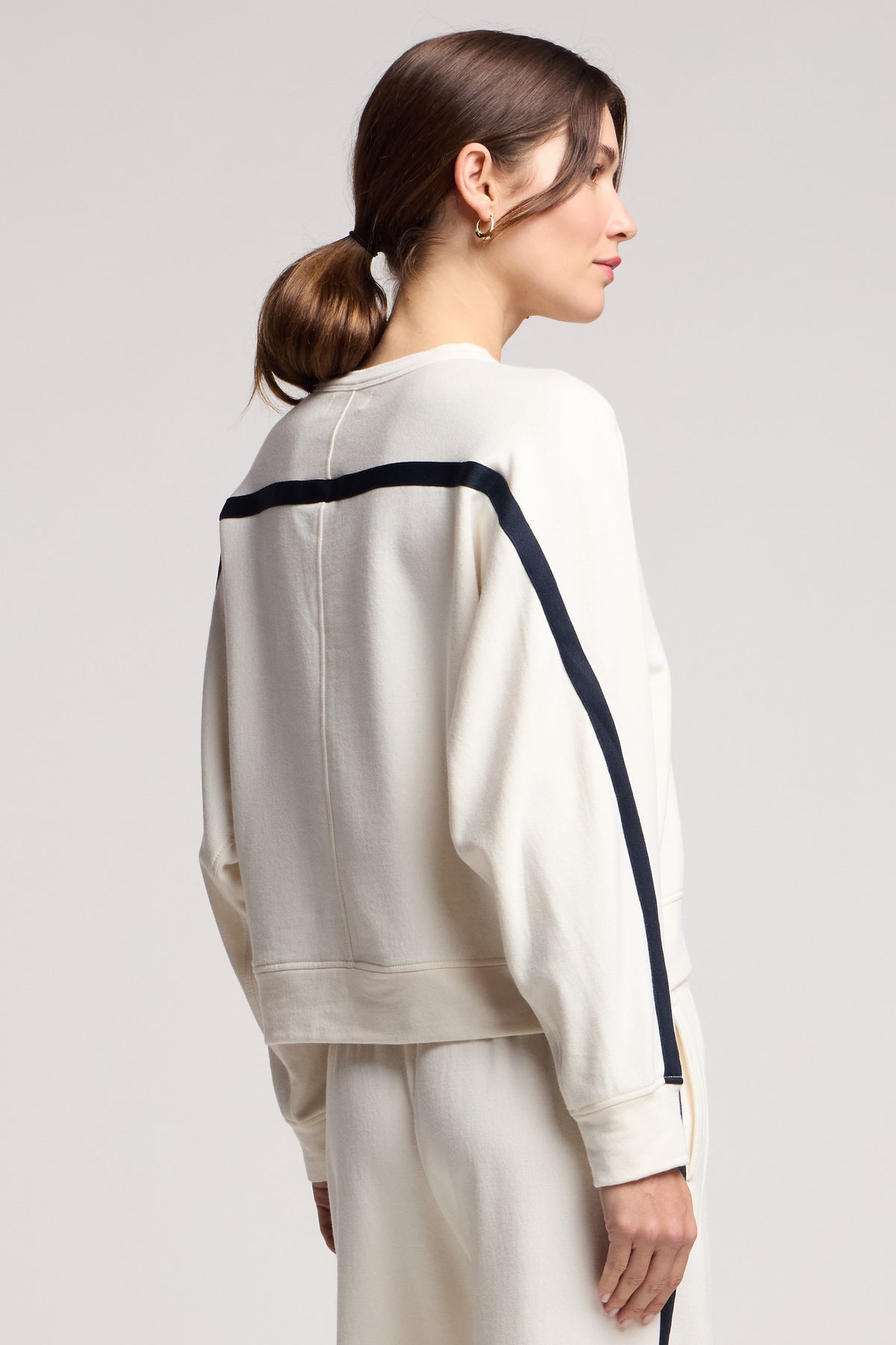 A woman with brown hair in a low ponytail wears the FRANKIE SWEATSHIRT by Velvet by Graham & Spencer—an off-white midweight fleece with a black stripe—paired with matching pants, facing sideways for a coordinated loungewear look.