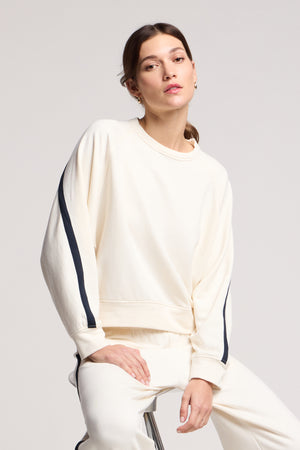 A person wears the FRANKIE SWEATSHIRT by Velvet by Graham & Spencer—cream-colored with black sleeve stripes—paired with matching pants, sitting on a stool and highlighting effortlessly coordinated loungewear.