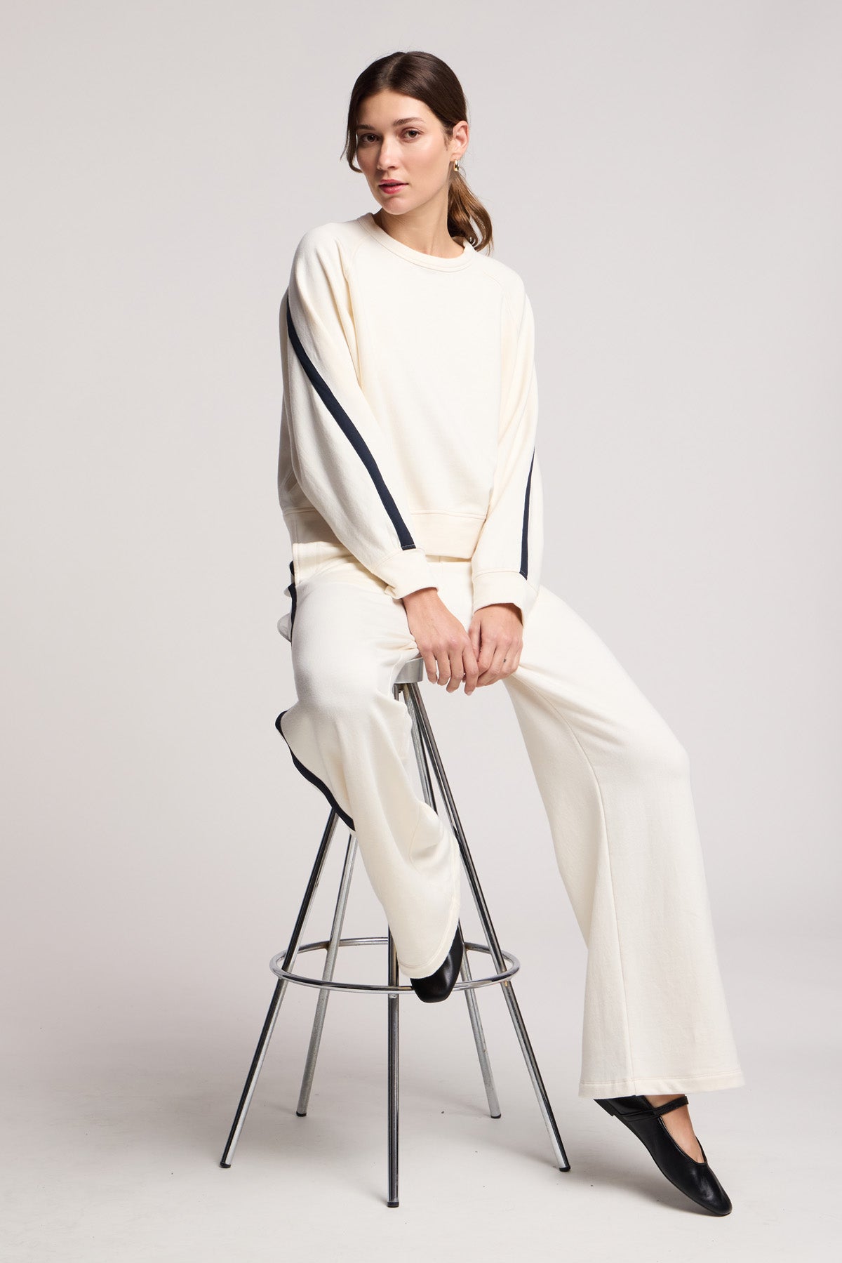 A woman sits on a tall metal stool against a plain background, wearing Velvet by Graham & Spencer’s cream-colored GIGI SWEATPANT with black stripe accents and a matching elastic waist sweatshirt.