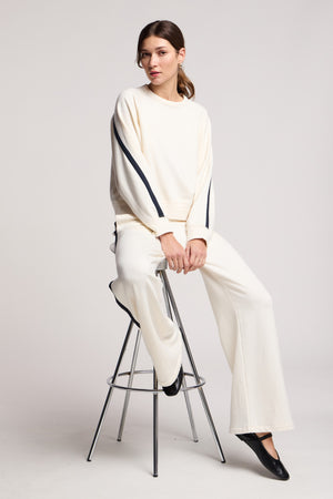 A woman sits on a tall metal stool against a plain background, wearing Velvet by Graham & Spencer’s cream-colored GIGI SWEATPANT with black stripe accents and a matching elastic waist sweatshirt.