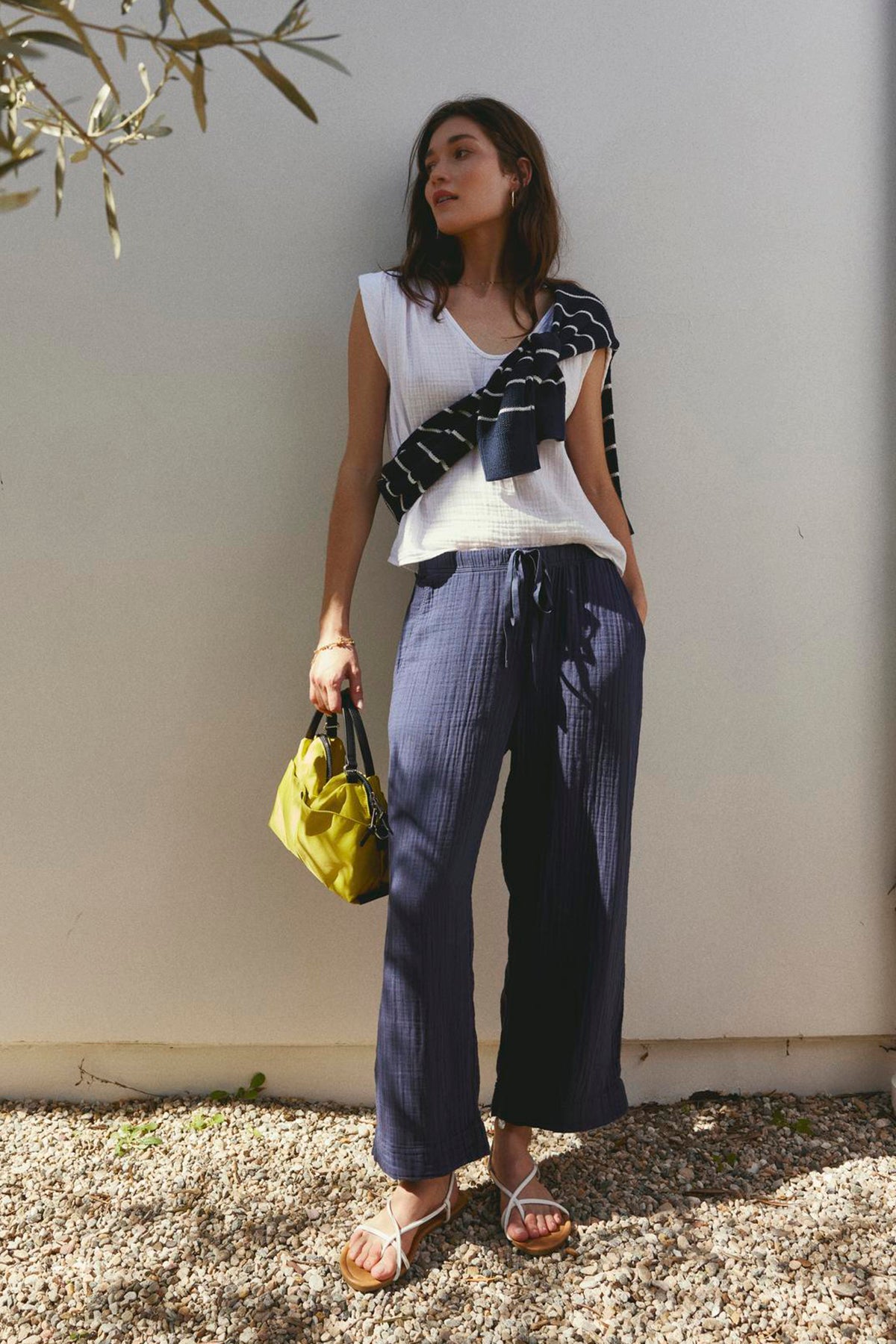   Woman standing in sunlight leaning against a wall, wearing a CHAYSE STRIPED CREW NECK SWEATER sweater from Velvet by Graham & Spencer, striped pants, and sandals, holding a green bag in a relaxed silhouette. 