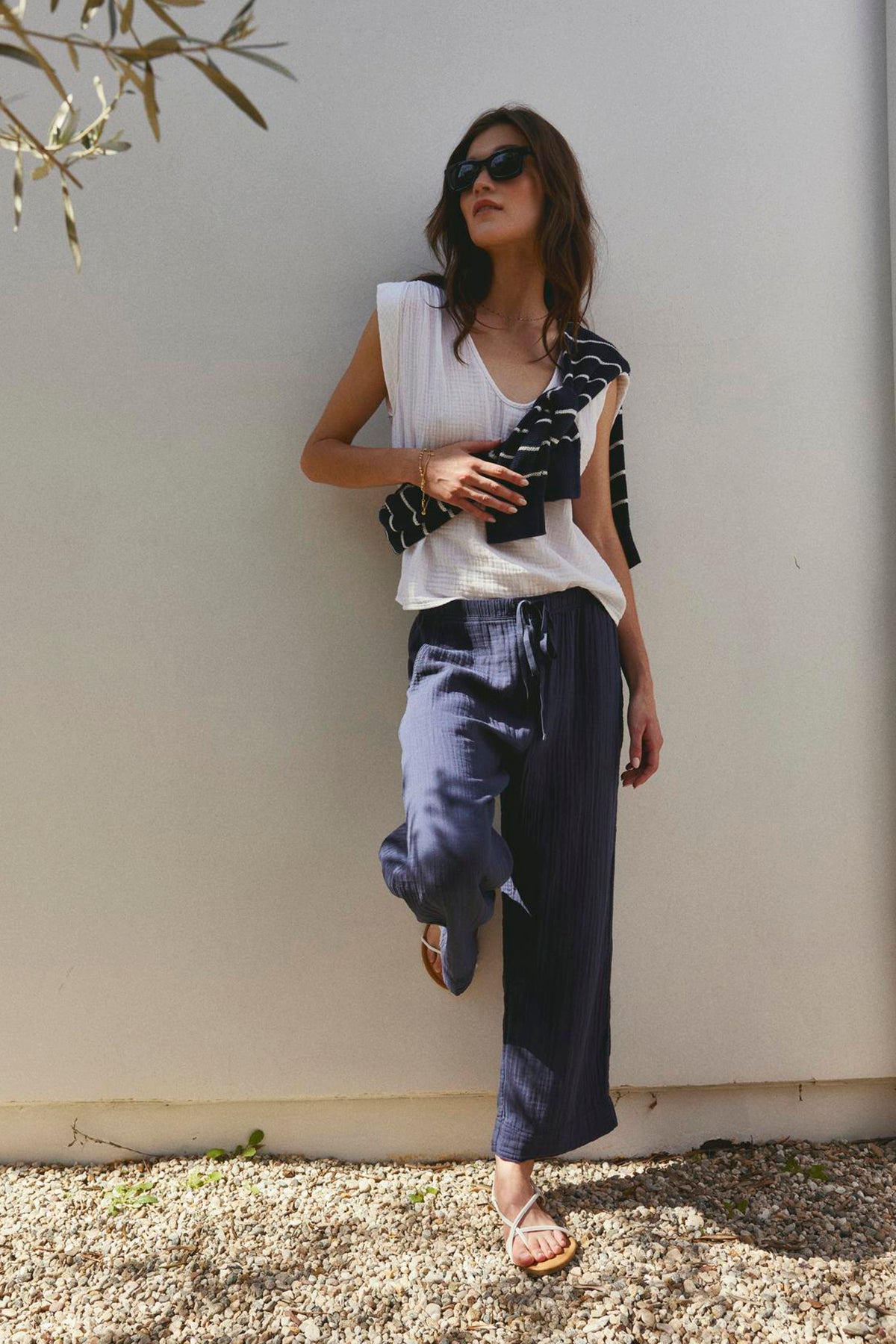   Woman in casual attire, wearing FRANNY COTTON GAUZE PANTS by Velvet by Graham & Spencer with slash pockets, leaning against a wall with sunglasses on. 