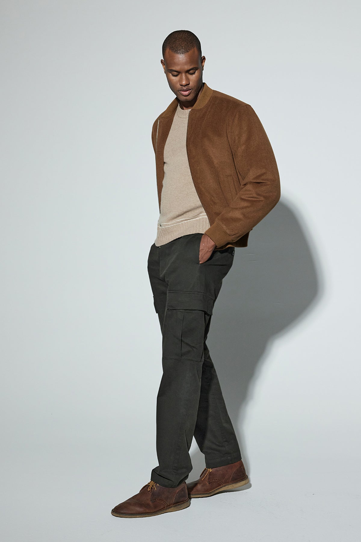   A person is standing with one hand in their pocket, wearing a brown jacket, beige sweater, BRYCE PANT mid-weight cotton lyocell twill dark green pants with an elasticized drawstring waist by Velvet by Graham & Spencer, and brown shoes. The background is plain and light-colored. 