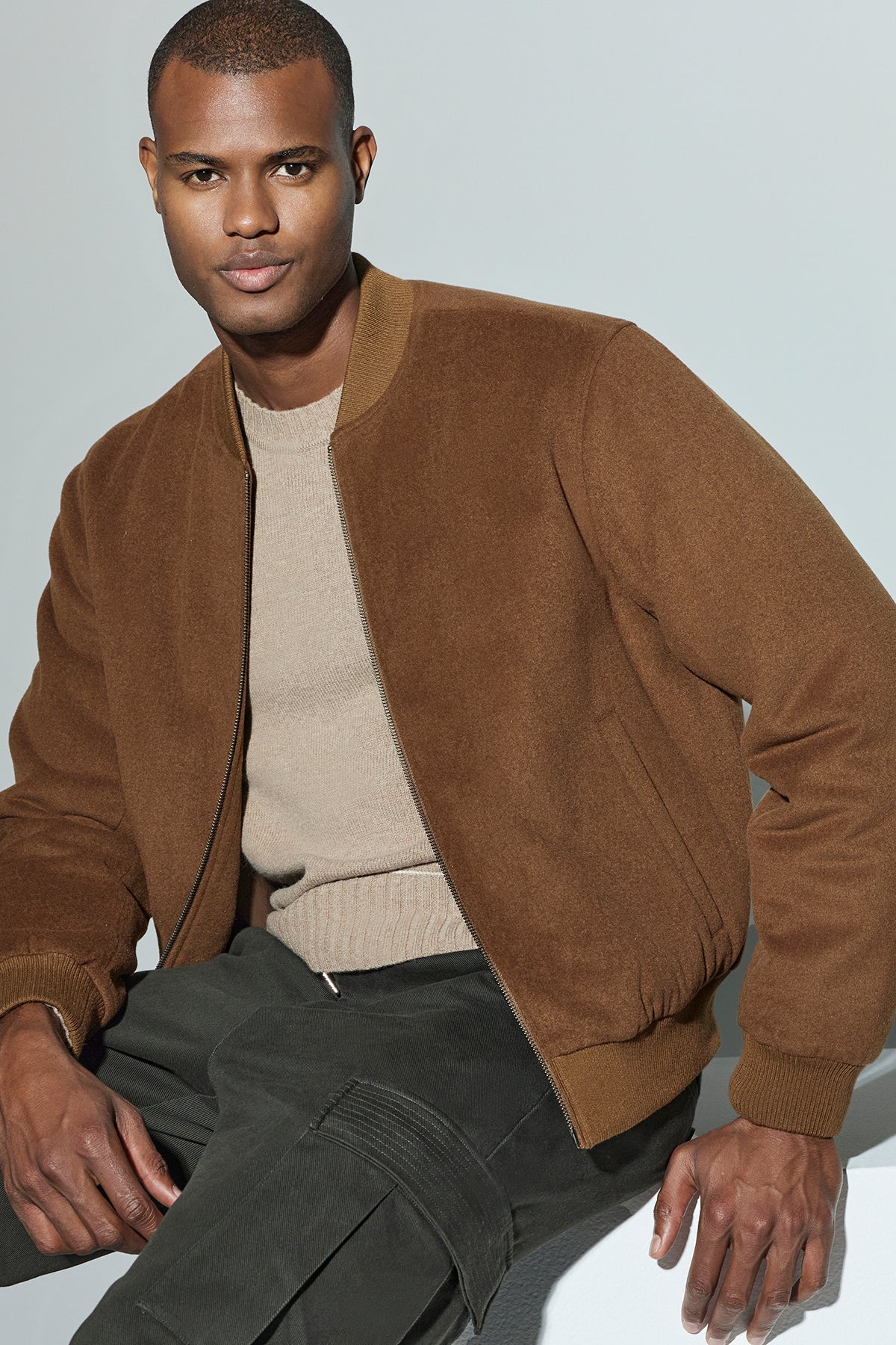 A man wearing the FRAZIER BOMBER JACKET by Velvet by Graham & Spencer and a beige sweater sits against a light background, exuding lasting warmth.-38218819502273