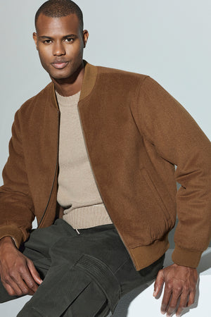 A man wearing the FRAZIER BOMBER JACKET by Velvet by Graham & Spencer and a beige sweater sits against a light background, exuding lasting warmth.